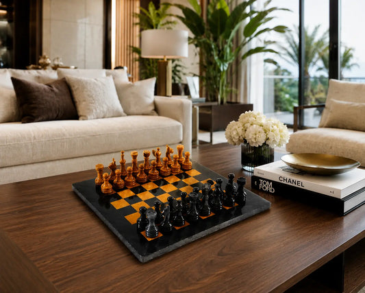 A luxury chess set with black and warm wood-toned pieces on a black slate board, styled on a dark wood coffee table in a modern, upscale living room with a beige sofa, plants, and decorative books.