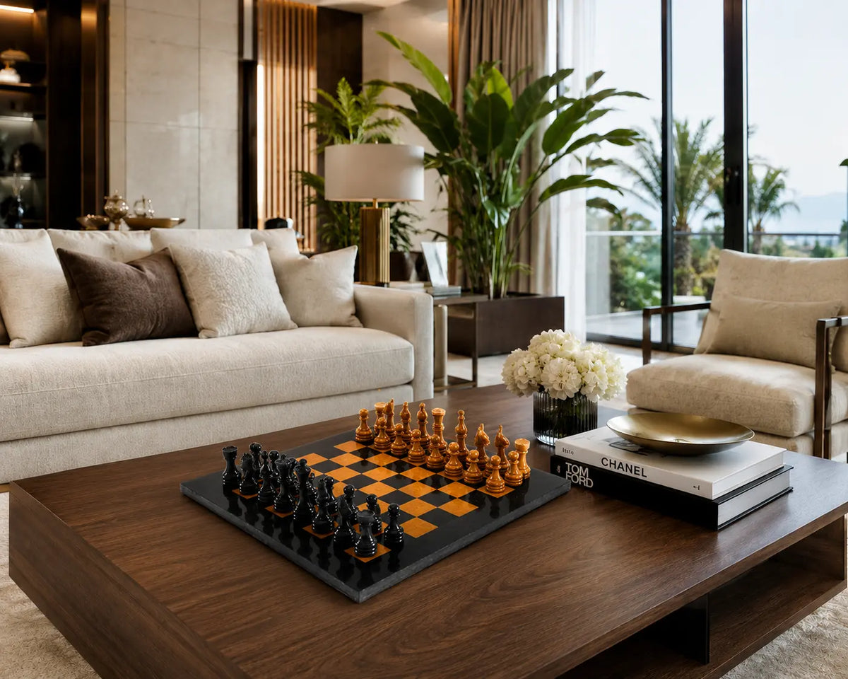A modern living room interior with a dark wood coffee table holding a black and orange chessboard and chess pieces, next to books and a vase of white flowers. The room features a cream sofa, a matching armchair, and large windows with a view of greenery outside.