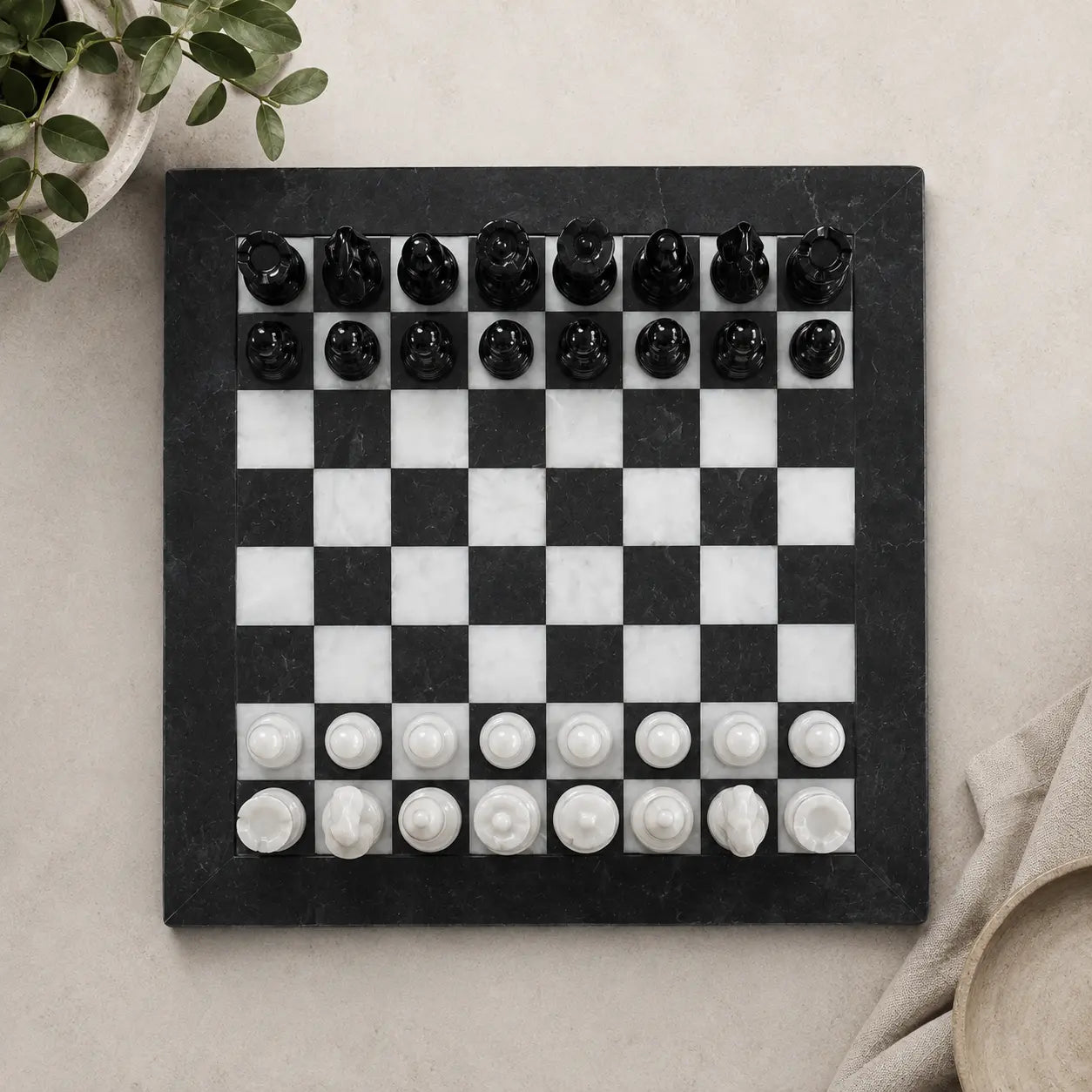 A black and white marble chessboard with carved black and white chess pieces, photographed from above on a light beige surface with small decorative plant leaves and fabric props at the edges.