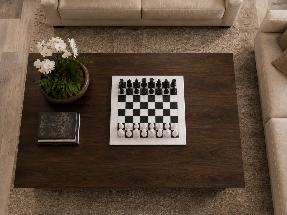 A top-down view of a dark wood coffee table in a living room setting, featuring a black and white chess set, a potted plant, and a book.