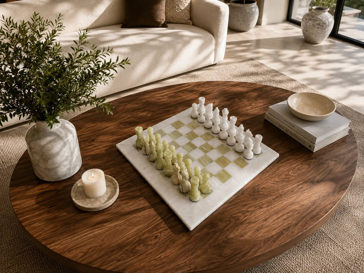 A marble chessboard with white and pale green chess pieces arranged on a round dark wood coffee table in a bright living room. A vase with greenery, a candle, books, and a bowl are also on the table.