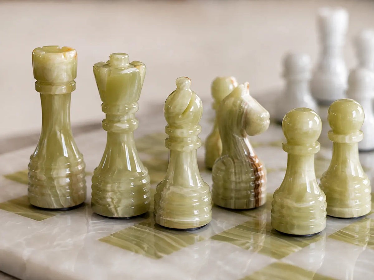 A set of polished onyx chess pieces in pale green and cream tones arranged on a matching marble chessboard, with a softly blurred background.